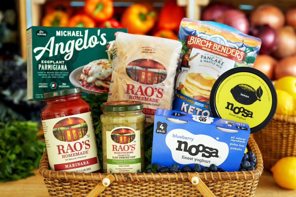 Basket containing various Sovos brand food products including Michael Angelo's Eggplant Parmigiana, Rao's Homemade Marinara Sauce, Italian Wedding Soup, Rigatoni Pasta, Birch Benders Pancake & Waffle Mix, and Noosa Lemon and Blueberry yoghurt.